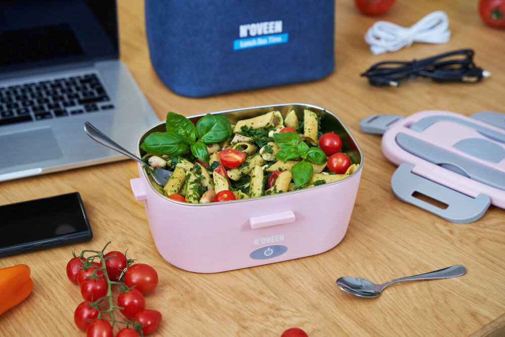 Na drewnianym stole znajduje się różowy elektryczny lunch box marki NOVEEN wypełniony makaronem z zielonymi liśćmi i pomidorkami. Obok leży pokrywka, łyżka, kabel oraz torba z napisem \'NOVEEN Lunch Box Store\'.