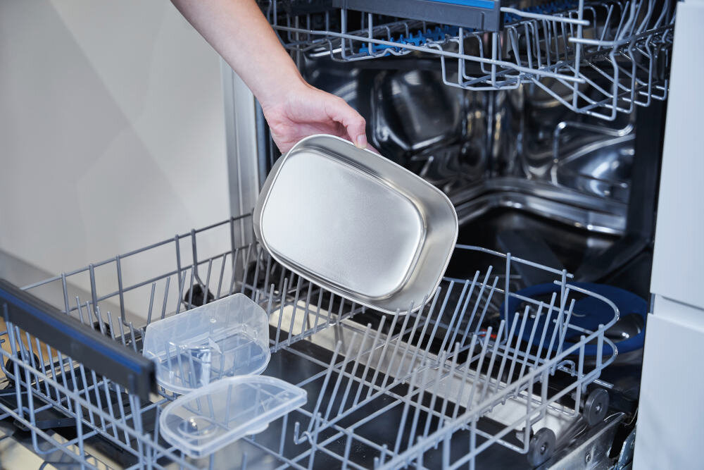 Osoba wkłada metalowy pojemnik do zmywarki, w której znajdują się również plastikowe elementy. Wnętrze zmywarki jest widoczne z otwartymi drzwiami.