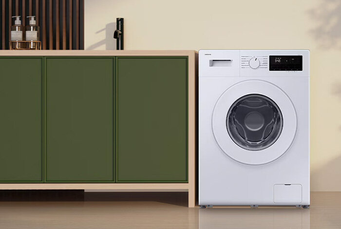 Pralka Slim 9kg WW90FG3M05TWEO Biała pralka z okrągłym, przezroczystym oknem znajduje się obok zielonej szafki. Na panelu pralki widoczny jest wyświetlacz i pokrętło do wyboru programów.