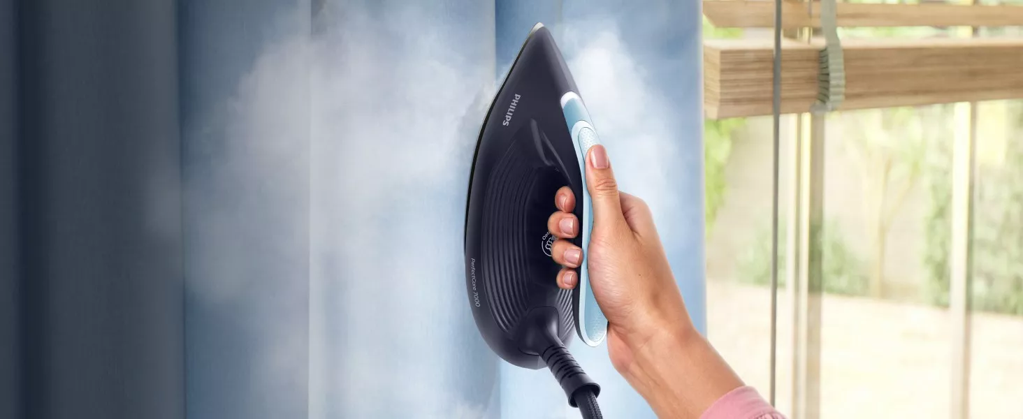 Osoba trzyma żelazko parowe marki PHILIPS, które emituje parę w kierunku niebieskiej tkaniny. Na żelazku widoczny jest napis \'PerfectCare 7000\'.