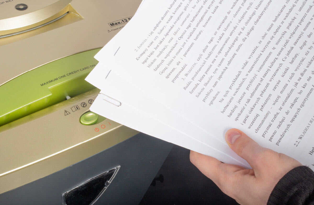 Osoba trzyma w ręku kilka zszytych kartek papieru nad niszczarką z widocznym napisem \'MAXIMUM ONE CREDIT CARD OR\'.