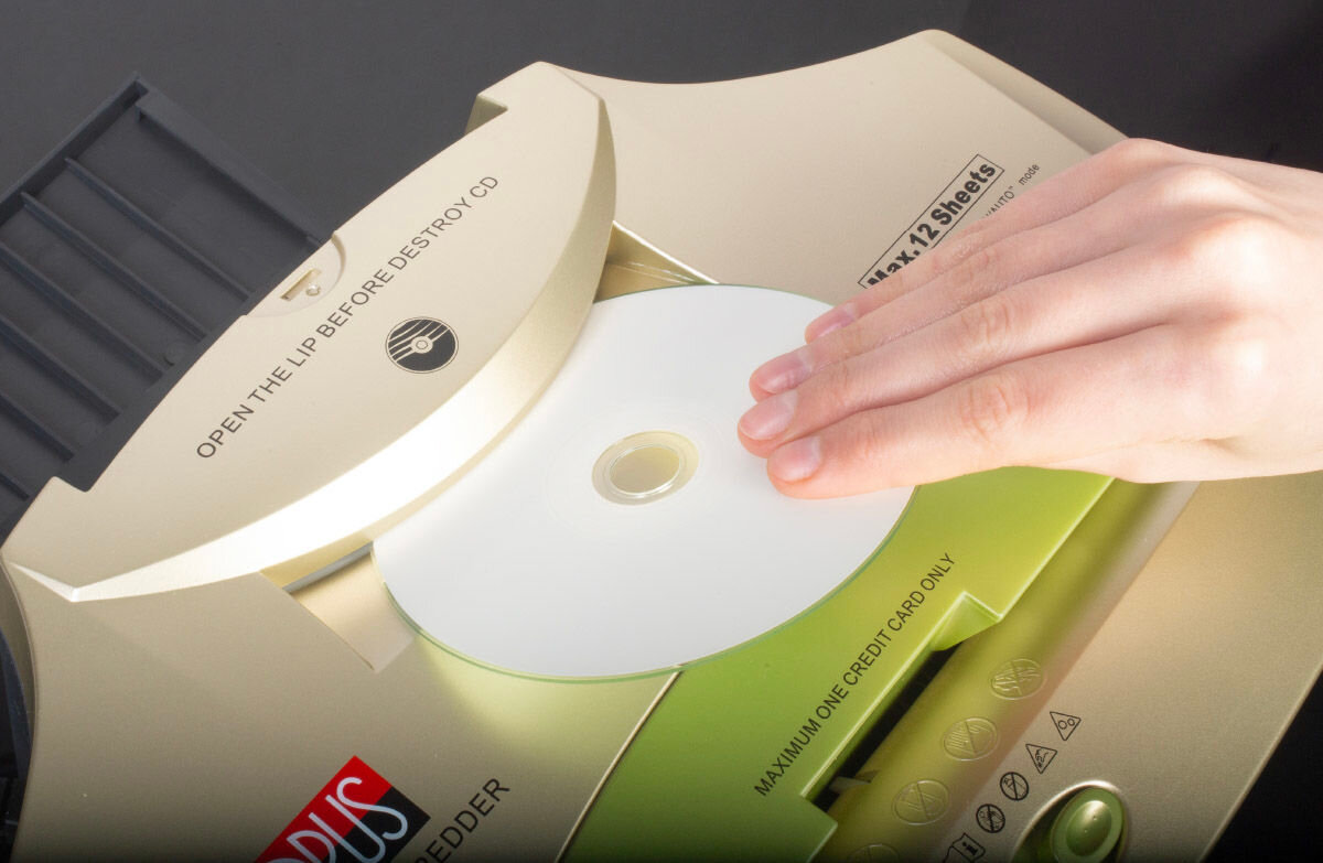 Osoba wkłada płytę CD do niszczarki w kolorze złotym z zielonym elementem. Na urządzeniu widoczny jest tekst: \'OPEN THE LIP BEFORE DESTROY CD\' oraz \'MAXIMUM ONE CREDIT CARD ONLY\'.