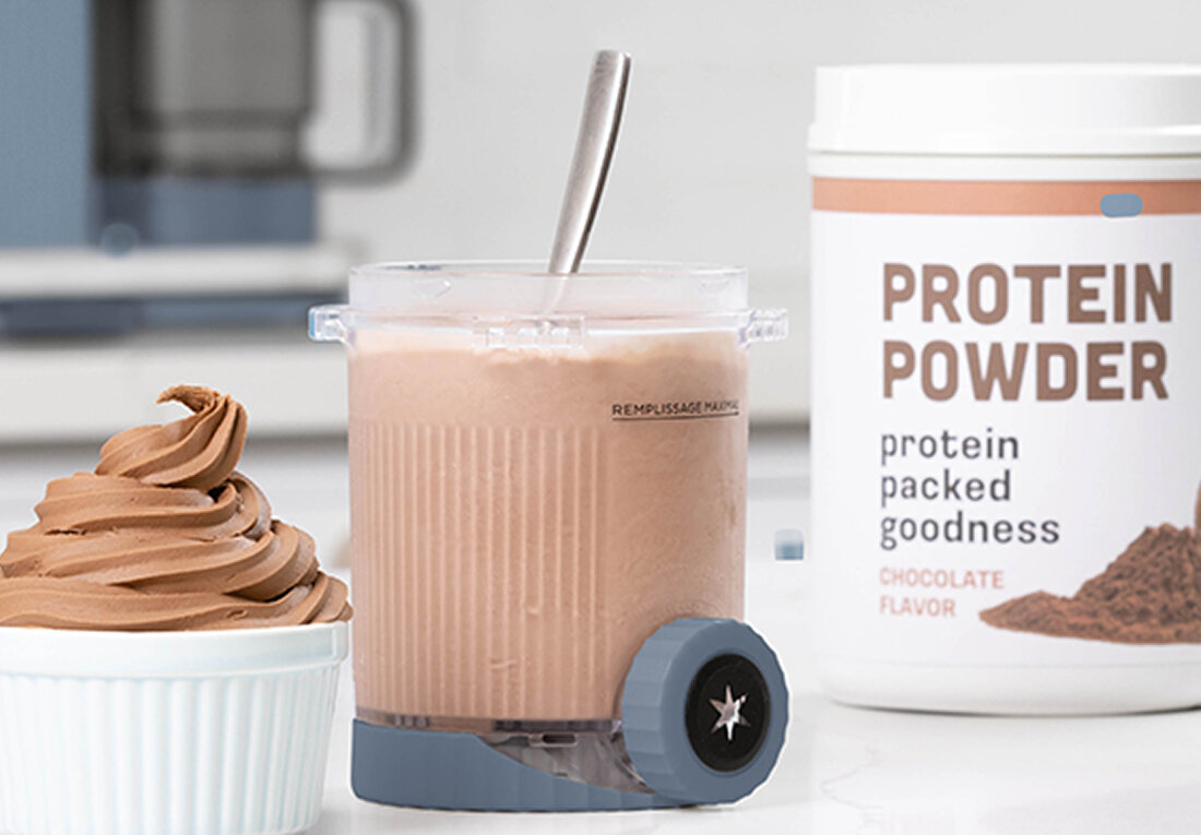 Na obrazie znajduje się pojemnik z różowym napojem i łyżką, obok miseczka z lodami oraz pojemnik z napisem \'PROTEIN POWDER protein packed goodness CHOCOLATE FLAVOR\'.