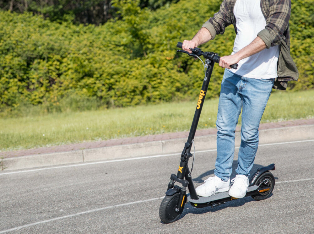 Osoba jedzie na czarnej hulajnodze elektrycznej z widocznym logo \'Jeep\' na kierownicy. W tle znajduje się zielony teren z krzewami.