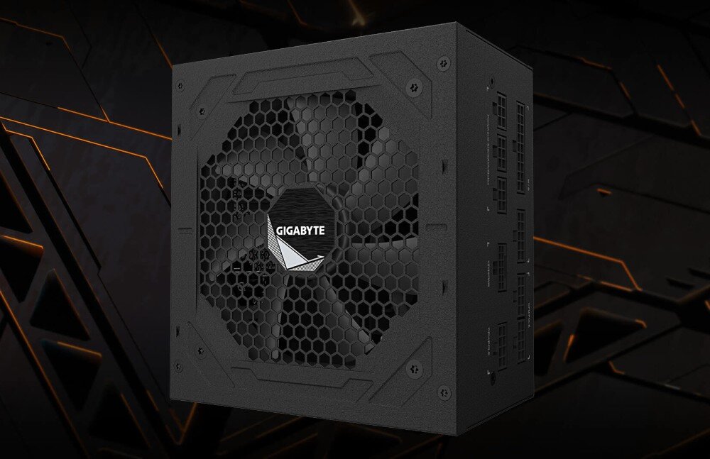 Na obrazie widoczny jest czarny zasilacz komputerowy z dużym wentylatorem i logo \'GIGABYTE\' na środku. Tło jest ciemne z pomarańczowymi liniami geometrycznymi.