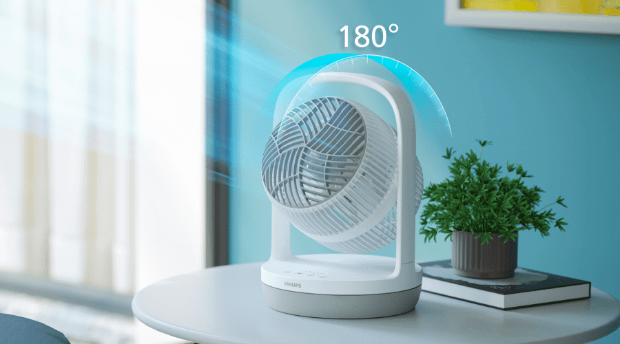 Na okrągłym stole stoi biały wentylator z logo PHILIPS, obok którego znajduje się zielona roślina w doniczce na książce. Nad wentylatorem widoczny jest napis \'180°\'.