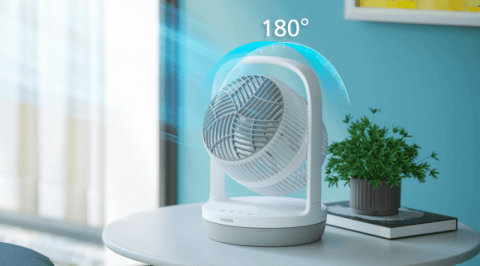 Na okrągłym stole stoi biały wentylator z napisem \'PHILIPS\' na podstawie, obok znajduje się zielona roślina w doniczce. Nad wentylatorem widoczny jest napis \'180°\'.
