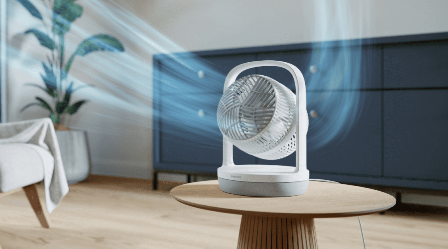 Na drewnianym stoliku stoi biały wentylator z logo PHILIPS, z którego wydobywają się niebieskie strumienie powietrza. W tle widać ciemną komodę i roślinę w doniczce.