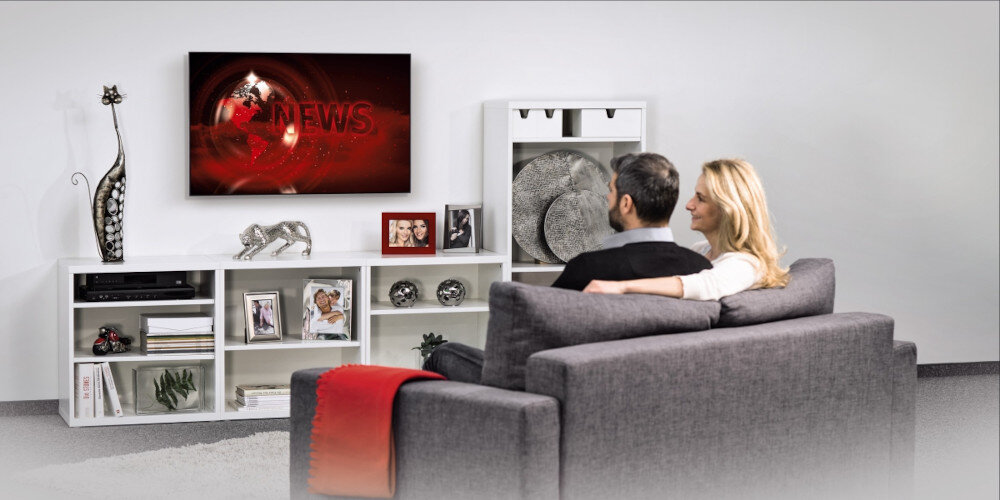 Para siedzi na szarej kanapie w salonie, oglądając telewizor zamontowany na ścianie. Na ekranie widoczny jest napis \'NEWS\', a wokół znajdują się białe półki z dekoracjami i zdjęciami.