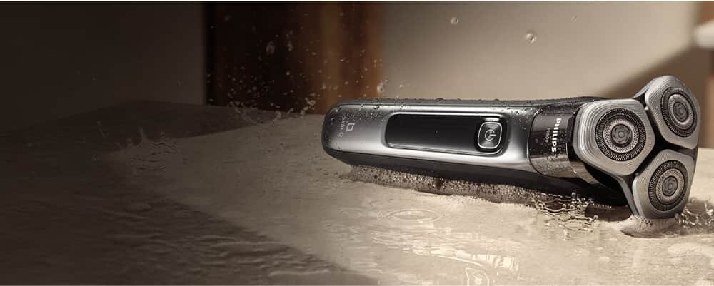 Golarka z trzema okrągłymi głowicami leży na mokrej powierzchni, z widocznymi kroplami wody. Na obudowie znajduje się logo PHILIPS.