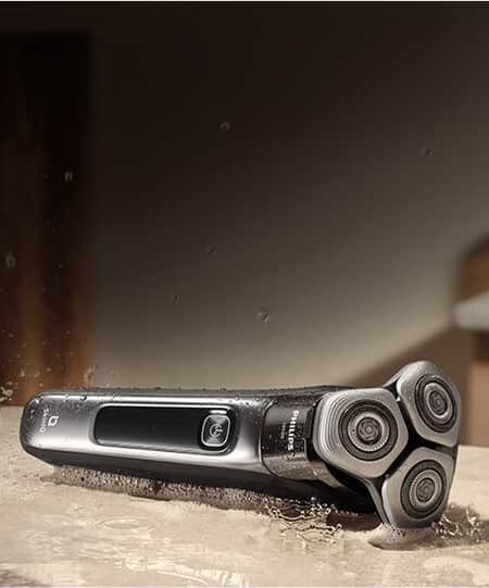 Na obrazie widoczna jest elektryczna golarka z trzema okrągłymi głowicami, leżąca na mokrej powierzchni. Na obudowie znajduje się logo \'PHILIPS\'.