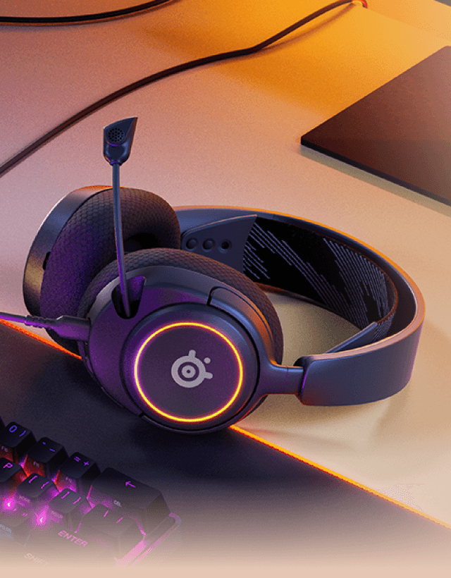 Czarne słuchawki nauszne z mikrofonem, z podświetleniem w kolorze pomarańczowym i logo SteelSeries na nauszniku. W tle widoczna jest klawiatura z podświetleniem oraz fragment biurka.