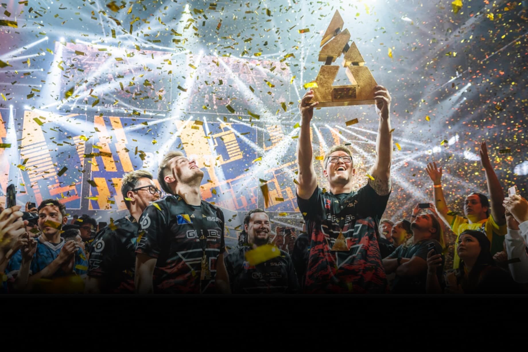 Grupa osób w koszulkach z logo G2 Esports świętuje, trzymając złote trofeum w otoczeniu konfetti i świateł scenicznych.