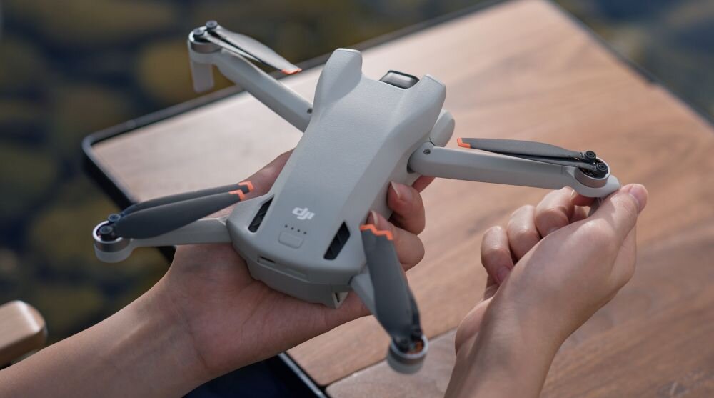 Osoba trzyma szary dron z rozłożonymi ramionami i śmigłami. Na dronie widoczne jest logo \'dji\'.