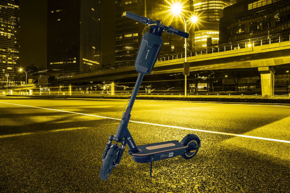 Czarna hulajnoga elektryczna XRIDER M10 Pro stoi na pustej, oświetlonej ulicy w mieście nocą. W tle widoczne są nowoczesne budynki i most.