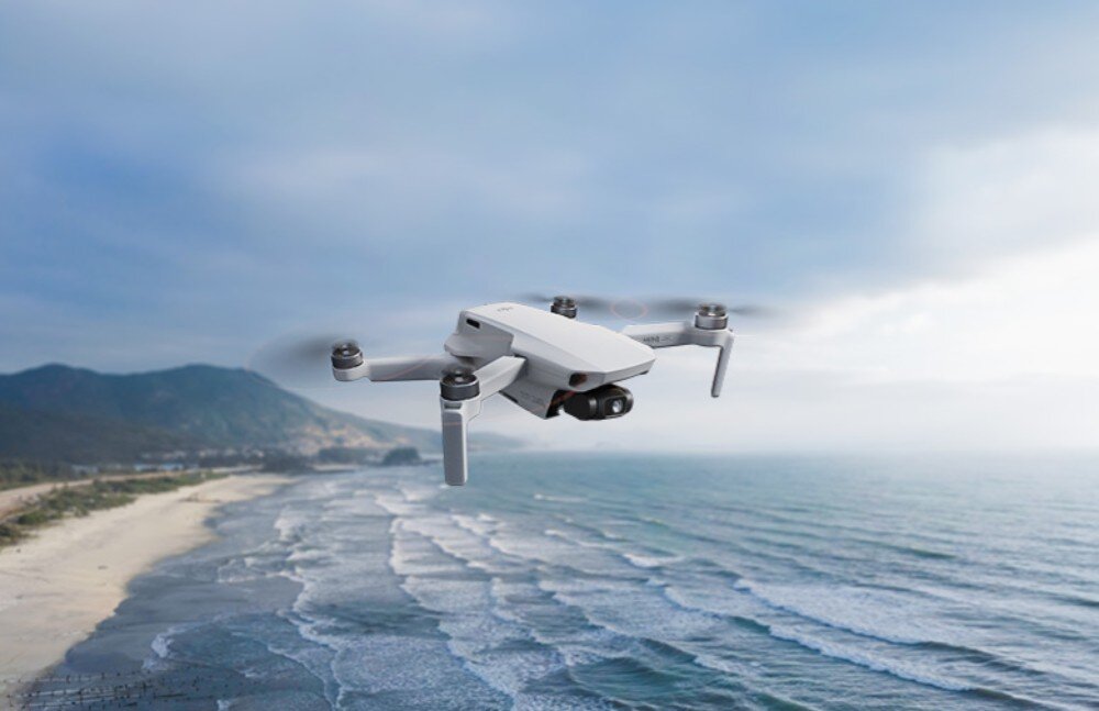 Dron unosi się nad linią brzegową z widokiem na morze i góry w tle. Niebo jest częściowo zachmurzone.