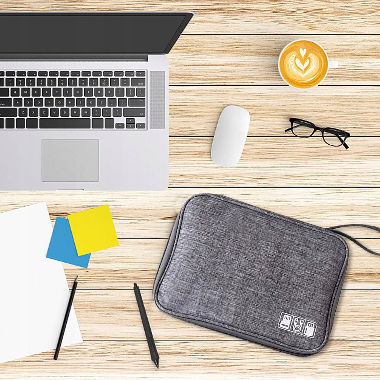 Na drewnianym blacie znajduje się otwarty laptop, mysz, filiżanka kawy z wzorem serca, okulary, dwa długopisy, kartki papieru, kolorowe karteczki oraz szara saszetka z symbolami kabli i ładowarek.