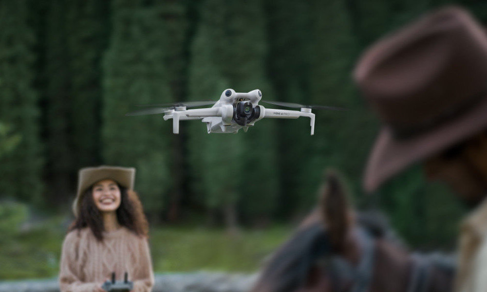 Dron unosi się w powietrzu na tle zielonego lasu. W tle widoczne są dwie osoby, jedna z nich trzyma kontroler.