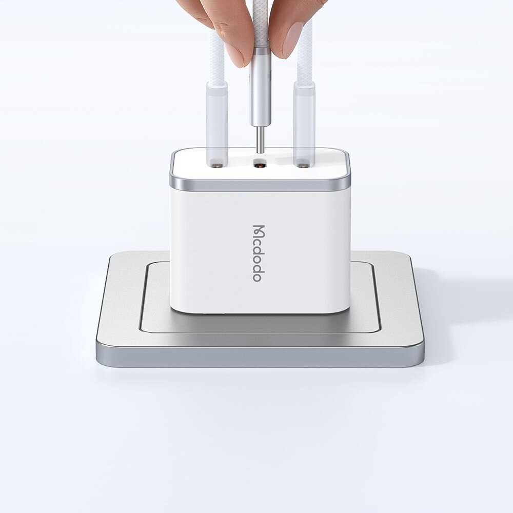 Biały adapter z logo \'Mcdodo\' na boku, z dwoma podłączonymi kablami USB-C, umieszczony na metalowej podstawce. Ręka trzyma jeden z kabli nad adapterem.