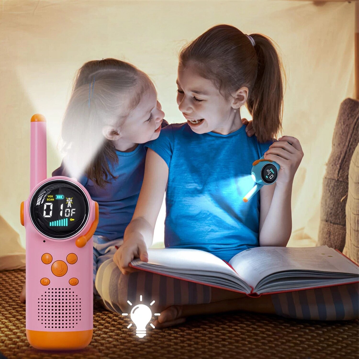 Dwoje dzieci siedzi w namiocie, jedno z nich trzyma otwartą książkę, a drugie trzyma walkie talkie z włączoną latarką. Obok widoczny jest różowy walkie talkie z wyświetlaczem pokazującym \'01\' i ikoną sygnału.