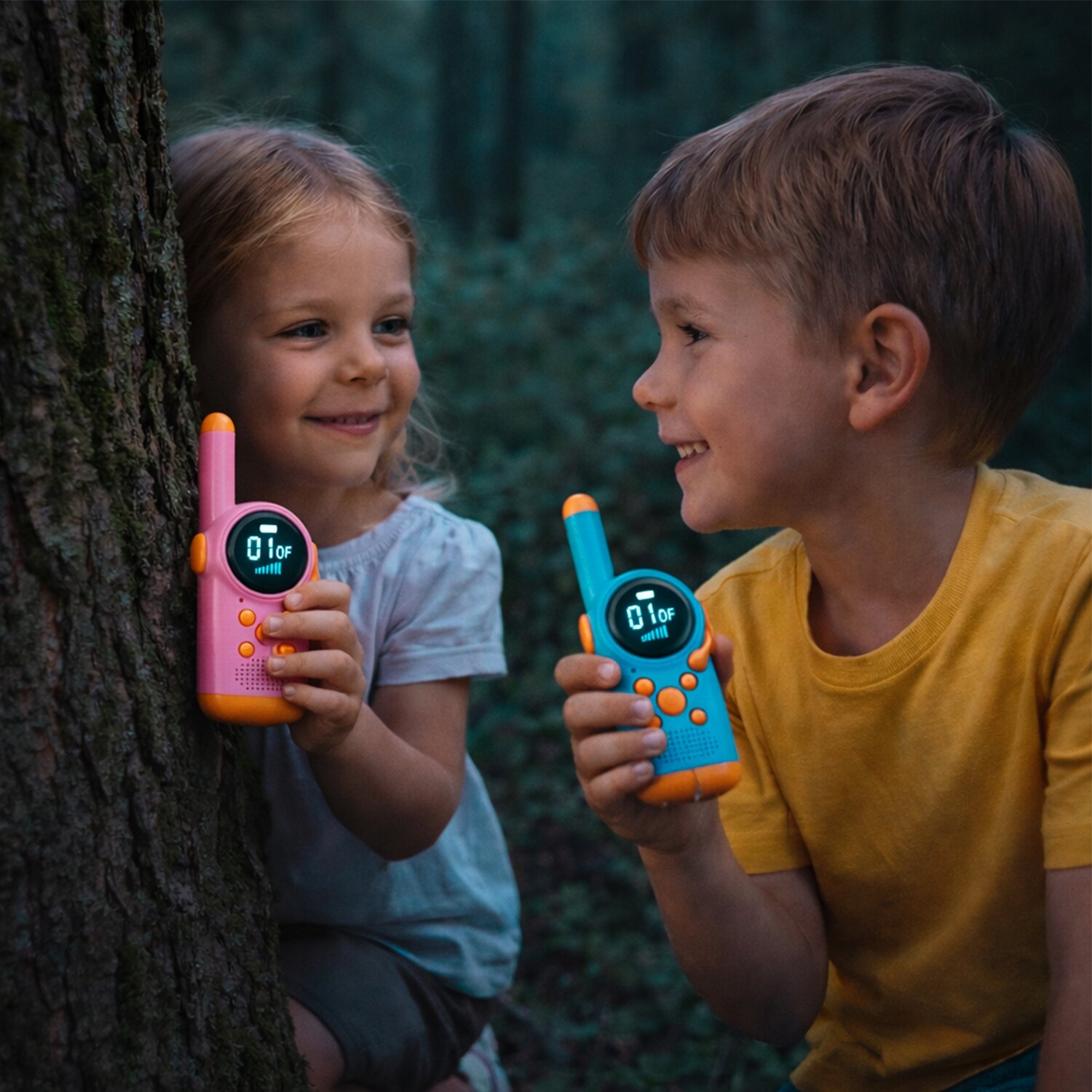 Dwoje dzieci trzyma kolorowe walkie talkie, jedno różowe, drugie niebieskie, z wyświetlaczem pokazującym \'01\' i ikoną sygnału. Dzieci są na zewnątrz, w tle widać drzewo i zieleń.