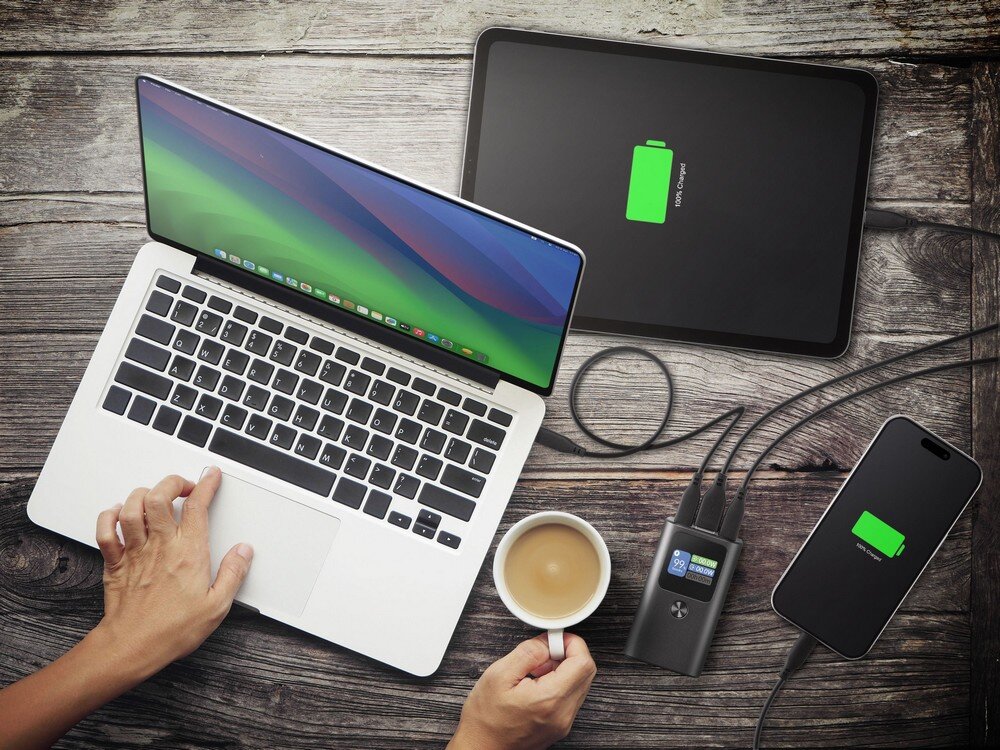 Na drewnianym stole znajduje się laptop, tablet i smartfon, wszystkie z ikoną ładowania baterii. Obok leży powerbank z wyświetlaczem pokazującym poziom naładowania, a obok niego filiżanka z napojem.