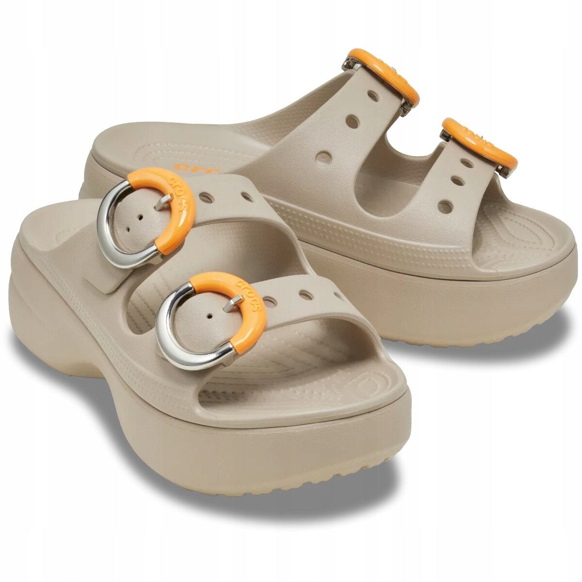Beżowe klapki na platformie z dwoma paskami, ozdobione okrągłymi klamrami w kolorze pomarańczowym i srebrnym. Na klamrach widoczny jest napis \'crocs\'.