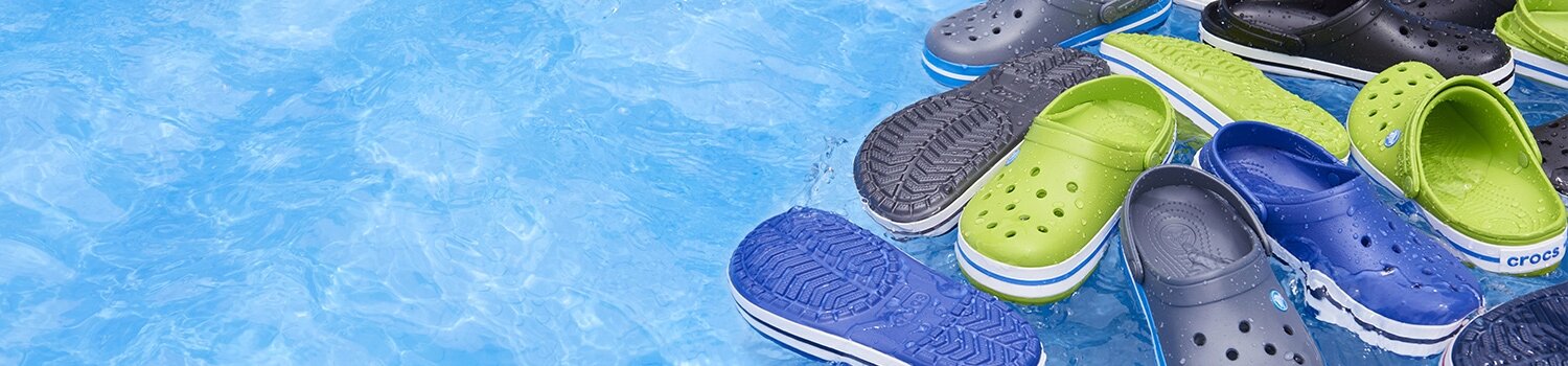 Na powierzchni wody unoszą się różnokolorowe chodaki, w tym zielone, niebieskie i czarne. Na jednym z chodaków widoczne jest logo \'crocs\'.