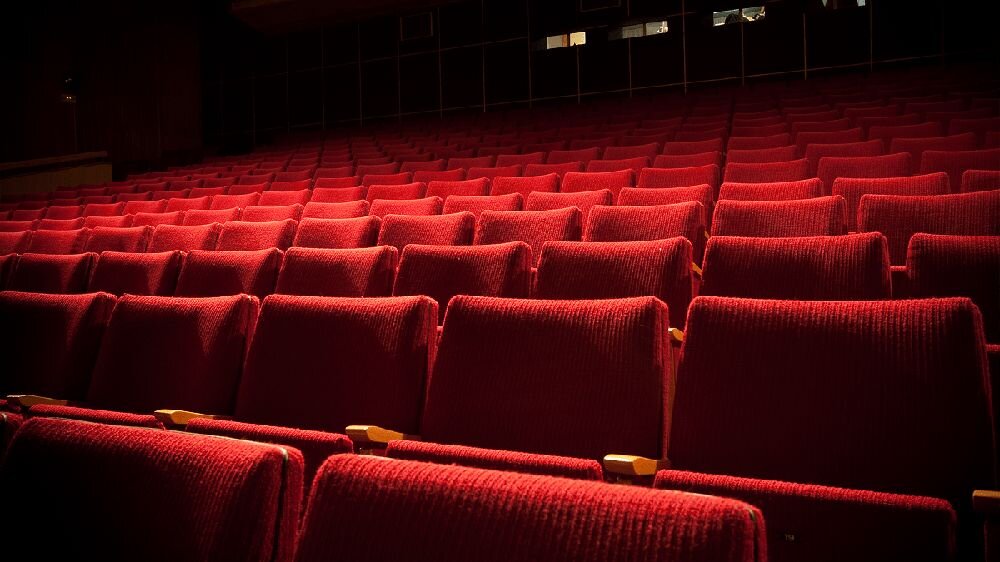 Zdjęcie przedstawia rzędy pustych, czerwonych foteli w sali kinowej lub teatralnej. W tle widoczne są ciemne ściany i niewielkie okna.