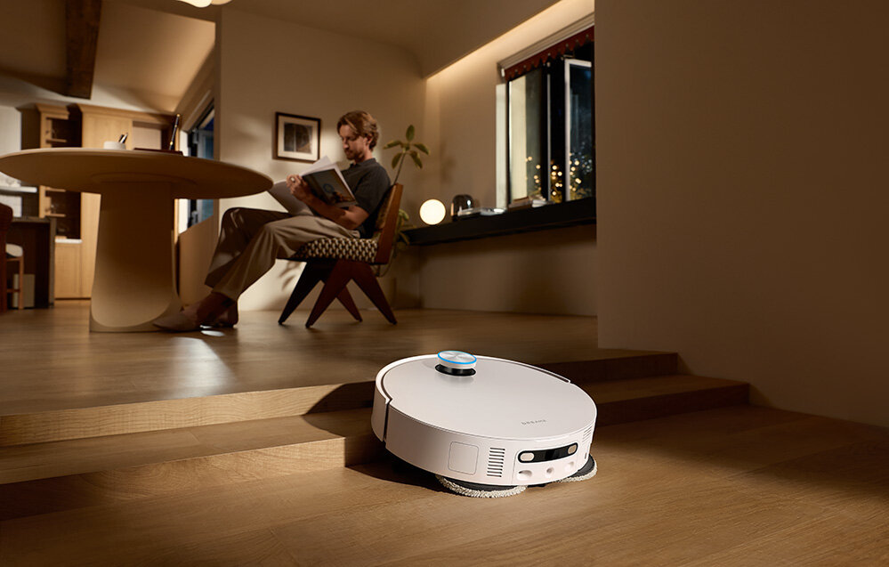 Na drewnianej podłodze znajduje się biały robot sprzątający. W tle osoba siedzi przy stole, czytając książkę.