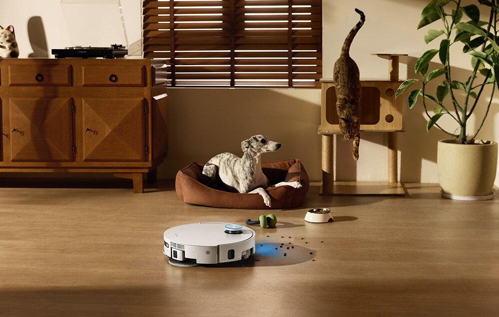 Na podłodze w salonie znajduje się biały robot sprzątający, wokół którego leżą rozsypane drobinki. W tle widać psa leżącego w legowisku oraz kota wspinającego się na drapak.