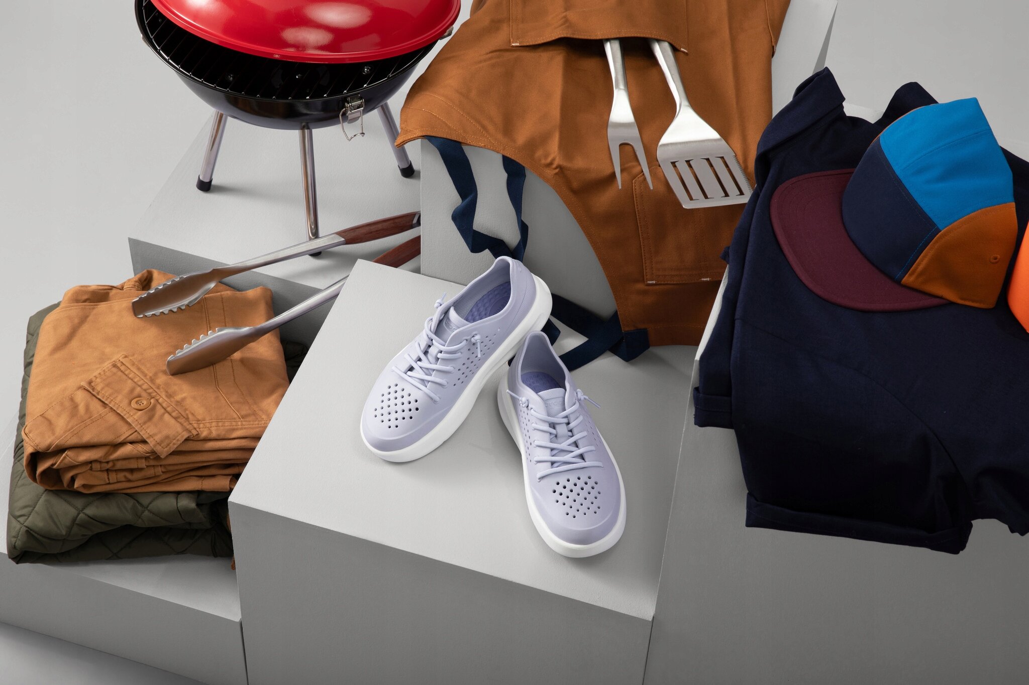 Na obrazie widoczne są białe buty sportowe, ubrania w różnych kolorach oraz akcesoria do grillowania, w tym czerwony grill i szczypce. Obok znajduje się czapka z daszkiem w kolorach granatowym, pomarańczowym i niebieskim.