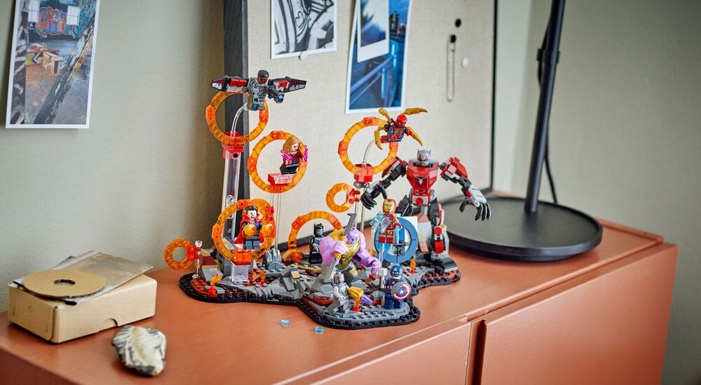 Na szafce znajduje się zestaw klocków LEGO przedstawiający scenę z figurkami superbohaterów i robotów. W tle widoczne są zdjęcia przypięte do tablicy oraz lampa biurkowa.