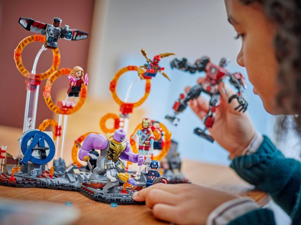 Dziecko bawi się zestawem klocków LEGO przedstawiającym scenę z postaciami superbohaterów i złoczyńców. W tle widoczne są konstrukcje z klocków z pomarańczowymi pierścieniami i figurki postaci w dynamicznych pozach.