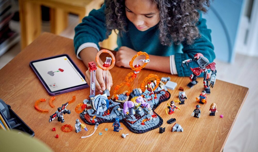 Dziecko bawi się zestawem klocków LEGO na drewnianym stole, obok leży tablet z instrukcją. Na stole znajdują się figurki i elementy konstrukcyjne związane z tematyką superbohaterów.
