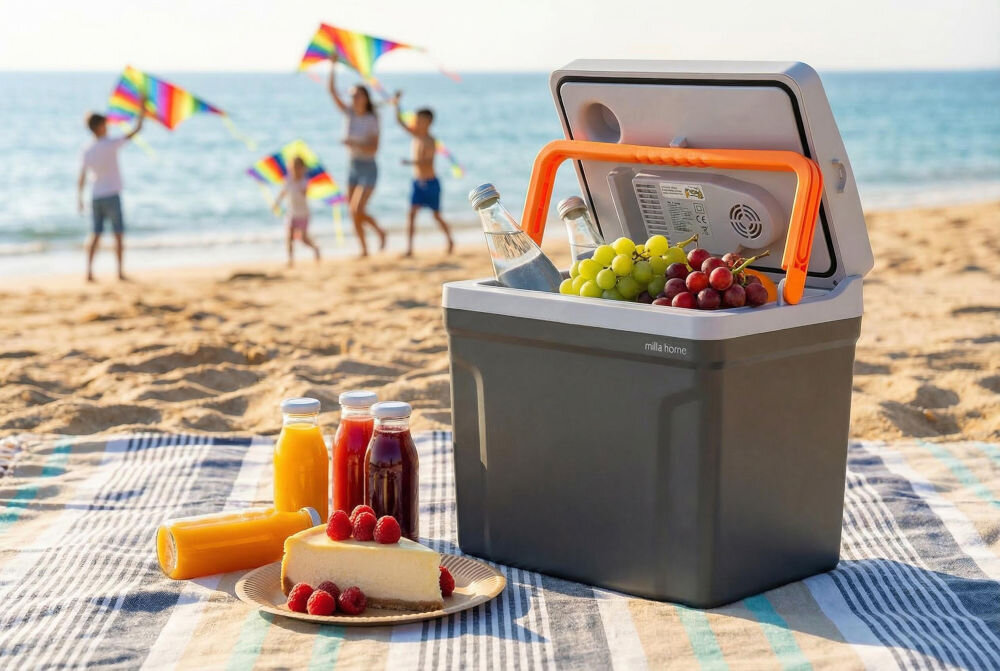 Na plaży stoi otwarta lodówka turystyczna z napisem \'milla home\', wypełniona butelkami i owocami. Na kocu obok znajdują się soki w butelkach i kawałek ciasta z malinami, a w tle widać ludzi z latawcami.