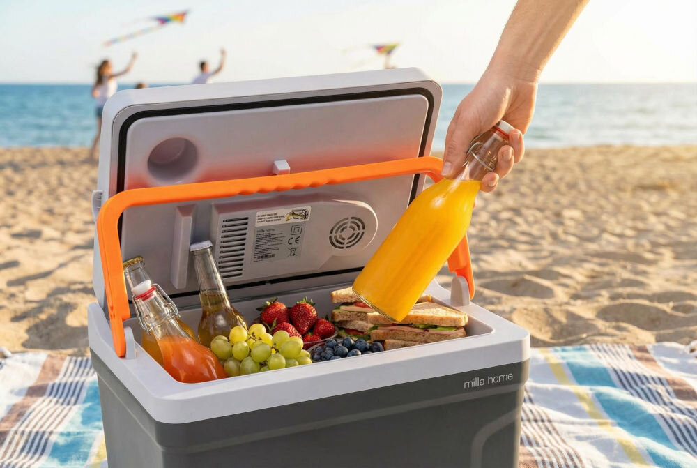 Na plaży znajduje się otwarta lodówka turystyczna marki \'milla home\' z różnymi produktami spożywczymi, takimi jak owoce i napoje. Osoba trzyma butelkę z pomarańczowym napojem.