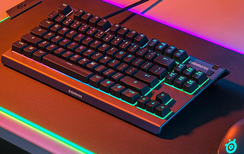 Klawiatura z podświetleniem RGB, widoczne logo \'steelseries\' na obudowie. Klawisze mają wyraźne oznaczenia, a całość znajduje się na podkładce z kolorowym podświetleniem.