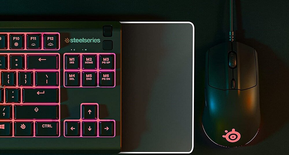 Na obrazie widoczna jest klawiatura z podświetleniem RGB oraz myszka z logo w kształcie okręgu i trzech kropek. Na klawiaturze znajduje się logo \'steelseries\'.