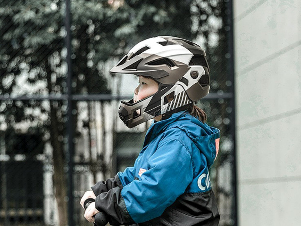 Dziecko w niebieskiej kurtce nosi szary kask rowerowy typu full face z odkręcaną szczęką. W tle widoczne są drzewa i ogrodzenie.