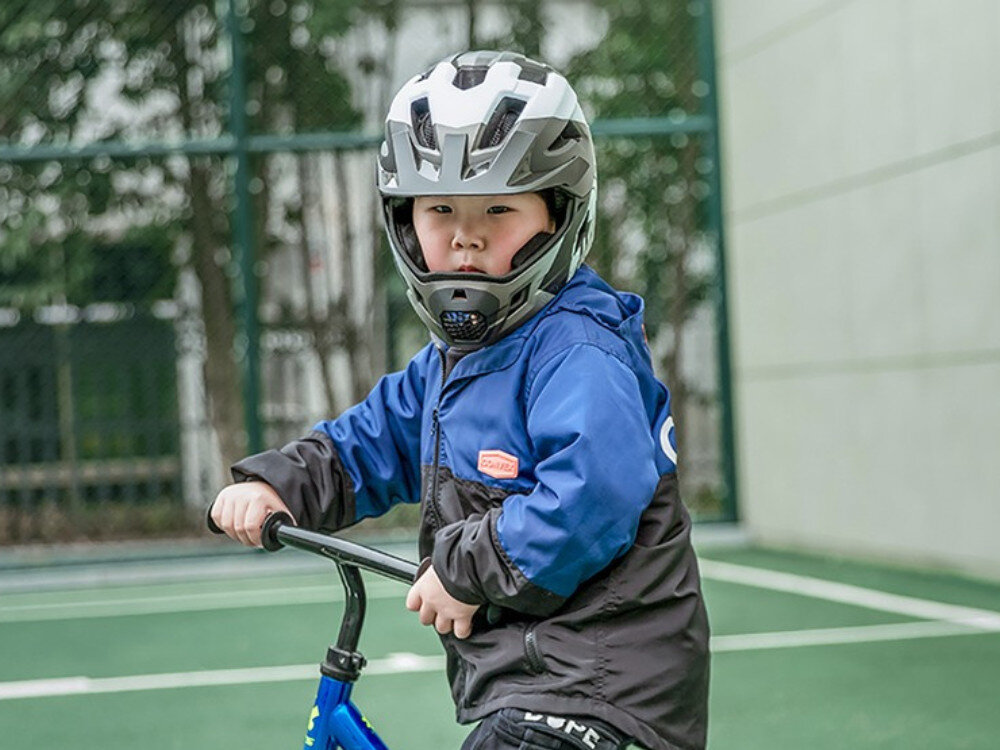 Dziecko w szarym kasku rowerowym z odkręcaną szczęką siedzi na rowerze na tle boiska sportowego. Kask ma nowoczesny design z wyraźnymi otworami wentylacyjnymi.