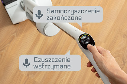Osoba trzyma odkurzacz z panelem sterowania, na którym widoczne są komunikaty: \'Samoczyszczenie zakończone\' i \'Czyszczenie wstrzymane\'.