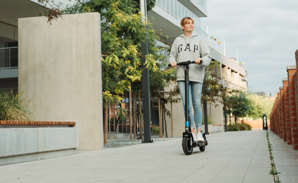Osoba jedzie na czarnej hulajnodze elektrycznej po chodniku w otoczeniu nowoczesnych budynków i zieleni. Na bluzie widoczny jest napis \'GAP\'.