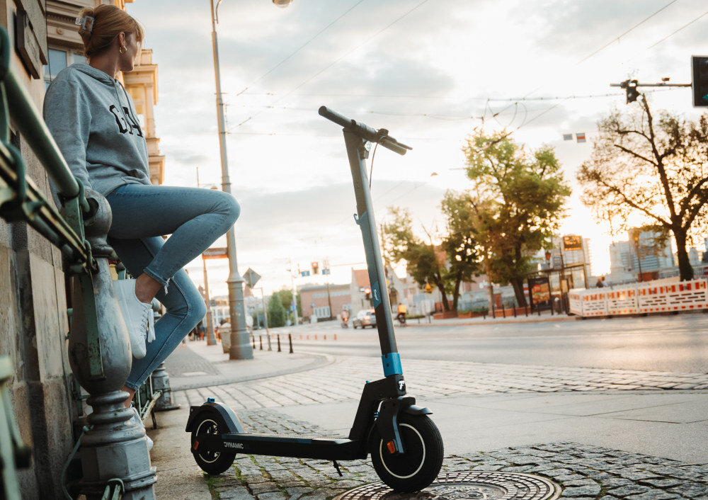 Na chodniku stoi czarna hulajnoga elektryczna z napisem \'DYNAMIC\' na boku. W tle widoczna jest osoba siedząca na murku oraz ulica z drzewami i budynkami.