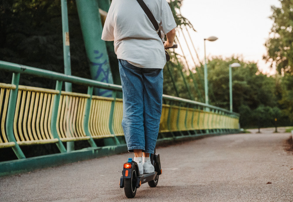 Osoba jedzie na czarnej hulajnodze elektrycznej po ścieżce na moście z zielonymi barierkami. W tle widać drzewa i latarnie.