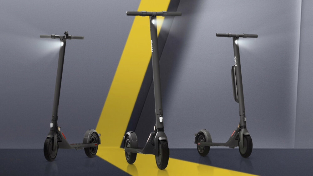 Trzy szare hulajnogi elektryczne ustawione na tle z żółtym wzorem. Na każdej hulajnodze widoczne jest logo \'segway\'.
