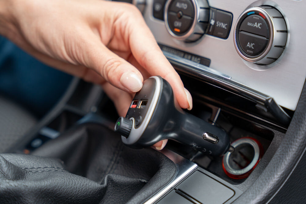 Osoba wkłada transmiter FM do gniazda zapalniczki w samochodzie. W tle widoczne są pokrętła klimatyzacji z oznaczeniami AUTO, SYNC, MAX A/C i A/C.