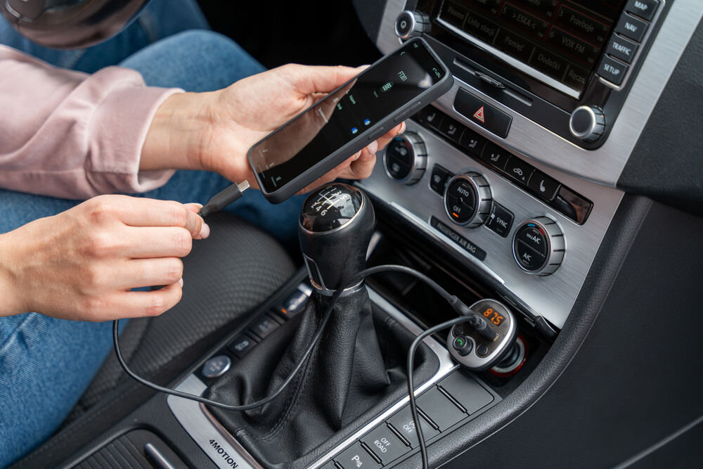 Osoba trzyma smartfon i podłącza go do transmitera FM w samochodzie. Na wyświetlaczu transmitera widoczna jest liczba \'87.5\'.