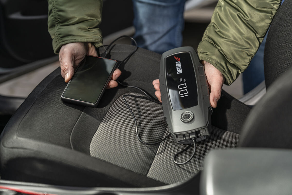 Osoba trzyma powerbank DEDRA z wyświetlaczem pokazującym \'100%\', podłączony do smartfona leżącego na siedzeniu samochodowym.