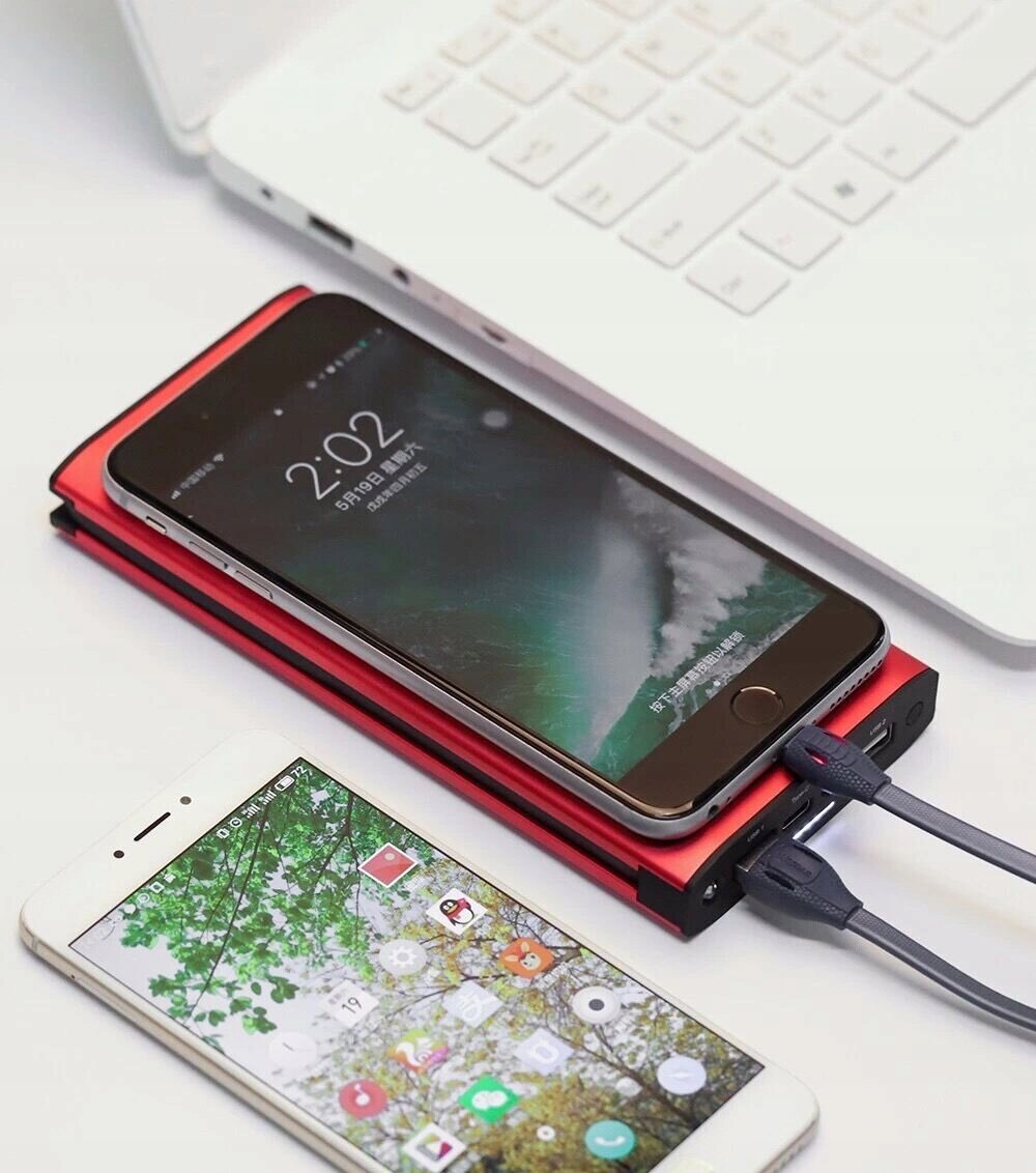Na obrazie widoczny jest aluminiowy powerbank z dwoma podłączonymi kablami, na którym leży smartfon z wyświetlaczem pokazującym godzinę 2:02. Obok znajduje się drugi smartfon z kolorowym ekranem, a w tle widać fragment białej klawiatury laptopa.